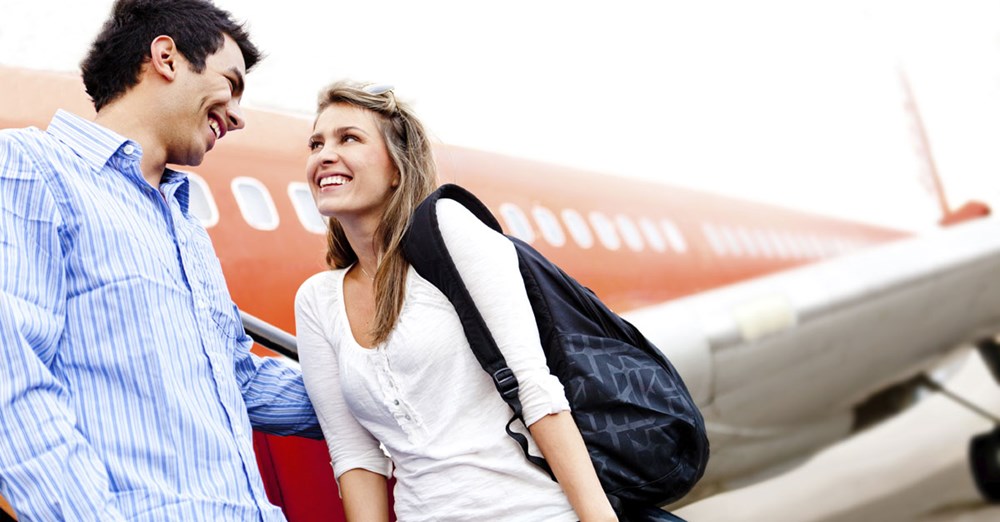 To smilende personer med ryggsekker ser hverandre foran et fly på en flyplass, de ser ut til å være glade og avslappede.